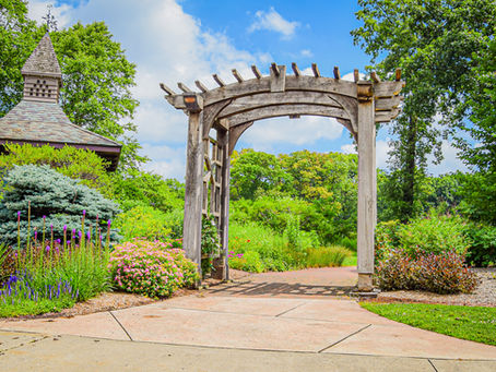 Wooden garden arch over a stone path beside colorful flowers and shrubs, with a small chapel roof and bright blue sky.
