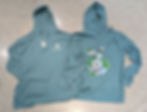 Two teal hoodies on a speckled floor. One hoodie shows a logo reading "Wellfield Botanic Gardens," the other features a flower and bee design.