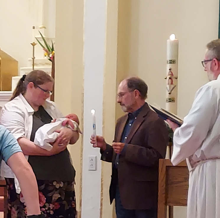 Child being held by his mother is presented with a baptismal candle by his godfather while priest watches