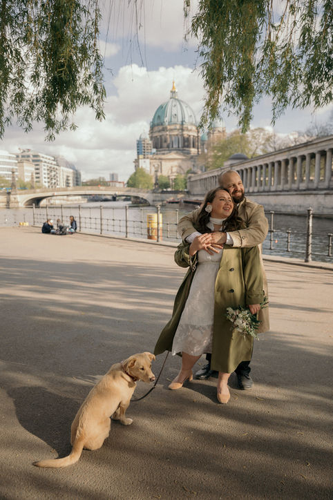 Die Braut und der Bräutigam umarmen sich, ihr Hund sitzt neben ihnen, der Berliner Dom im Hintergrund.