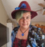 Person with green-tinted hair wearing a burgundy hat and floral jacket, smiling in a cozy room with a ceiling fan. Blue heart necklace.