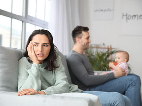 New parents sit on gray couch with baby. The father and playfully holds the baby atop his lap in backgrond while mother in is in foreground appearing distant and despondent. 