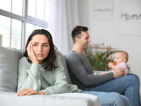 New parents sit on gray couch with baby. The father and playfully holds the baby atop his lap in backgrond while mother in is in foreground appearing distant and despondent. 