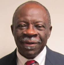 Smiling man in a suit and tie against a plain background. He has gray hair and a mustache. The mood appears warm and welcoming.