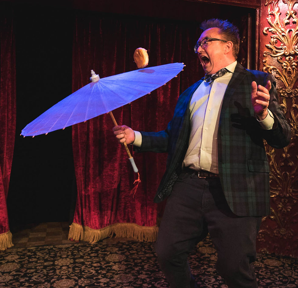 Performer on stage holding a large umbrella with a bagel balanced on top, delivering a lively and humorous variety act at a Laughter for All event.