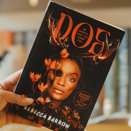 DOE by Rebecca Barrow being held by a brown hand in front of a book shelf