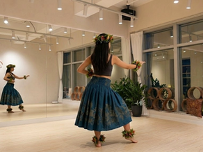A dancer practicing Polynesian Hula at Jiya Dance studio in Hong Kong. Wearing a teal botanical-patterned skirt and traditional floral lei accessories, she demonstrates a grounded posture and expressive storytelling hand movements reflected in the studio mirrors.