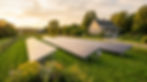 Solar panels in a grassy rural yard beside a farmhouse at sunset, with trees, fence, and wildflowers.