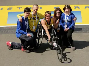 Athlete in a wheelchair racer, flanked by four supporters wearing blue MR8 shirts, posing on Boston Marathon finish line. Smiling, joyful scene.
