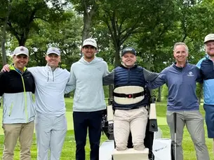 Eight men pose smiling on a green golf course, arms around each other, with trees and a golf cart behind them.
