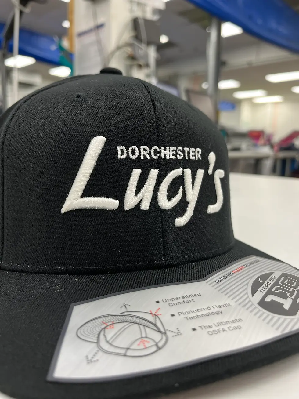 Black cap with "Dorchester Lucy’s" in white text, set on a table. Background shows a room with blue accents and lights. Emphasis on label details.