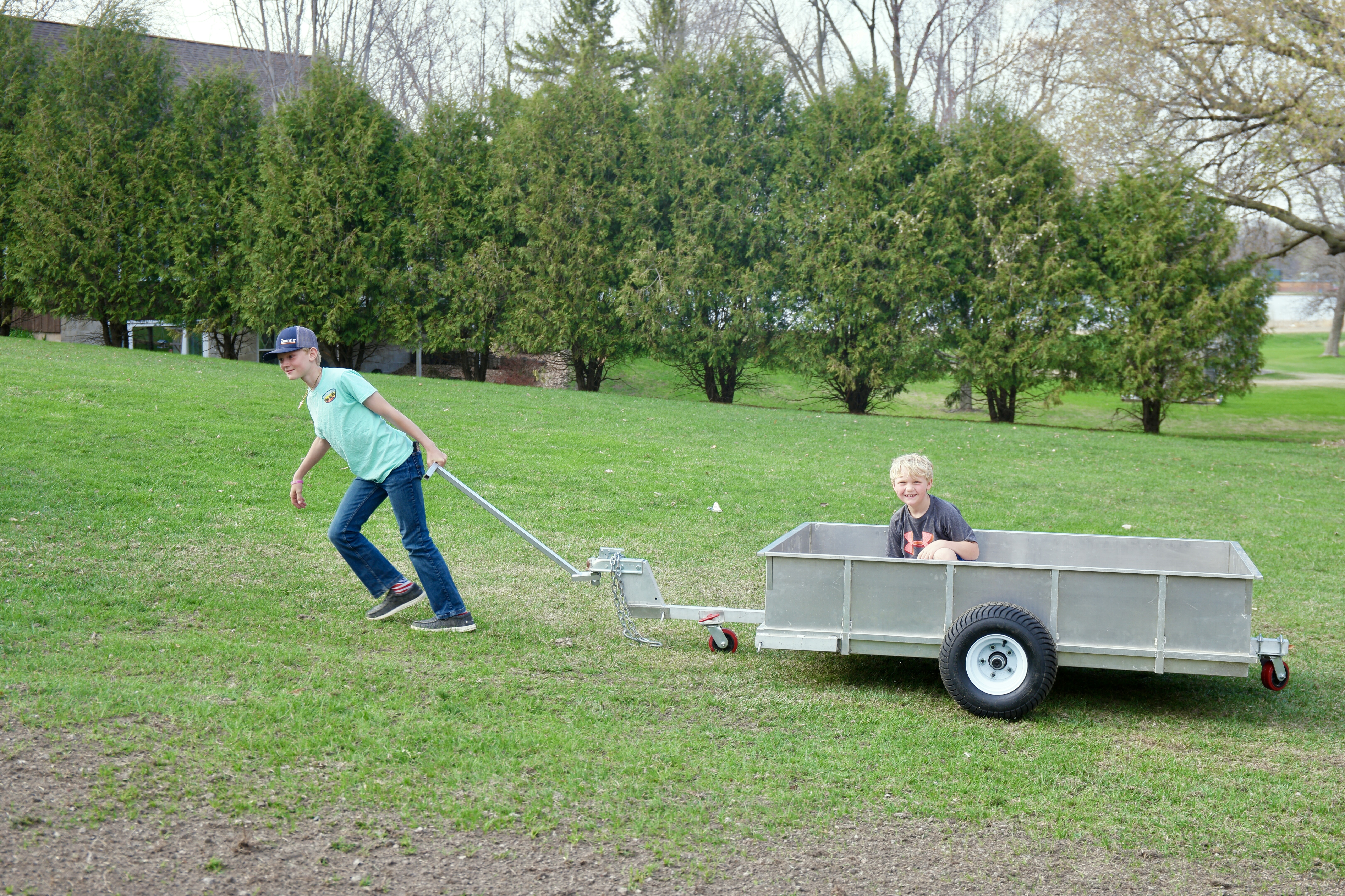 Two children in a yard with a Rambo Trailer, an aluminium multi-use on and off road trailer. One child is pulling the trailer with a handle, one child is riding in the bucket. 