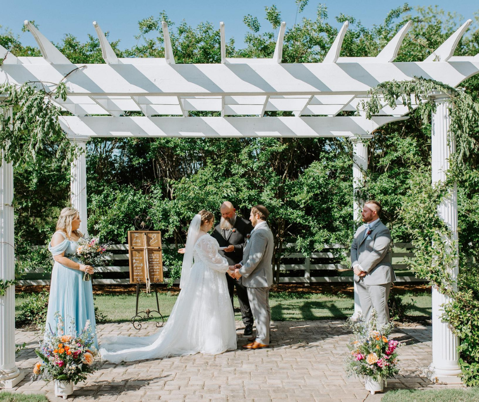 Bride and groom holding hands under a white pergola, with bridesmaid in blue and groomsman. Lush greenery, sunny day, outdoor ceremony.