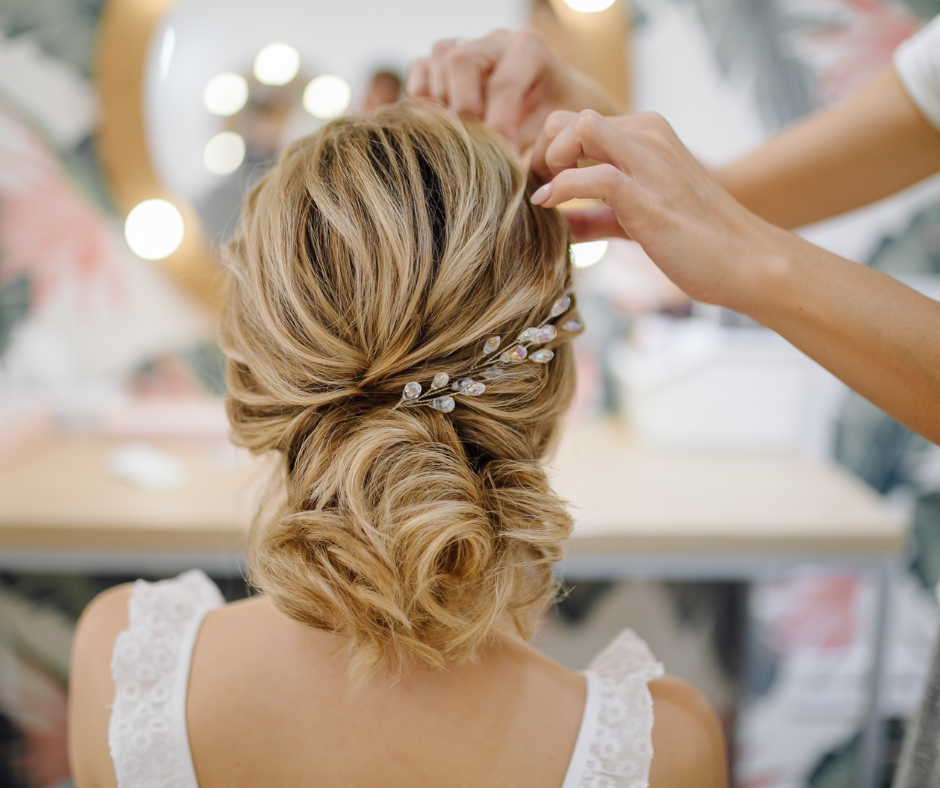 Stylist arranges a woman's blonde hair into an elegant updo with jeweled clips. Bright, soft-focus background with floral patterns.
