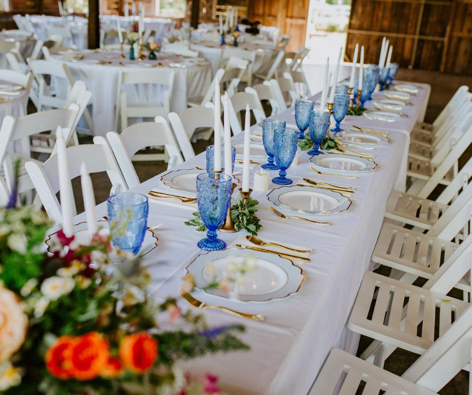 Elegant table setup with blue glasses, white plates, and candles on a white tablecloth. Floral decor and white chairs in a rustic setting.