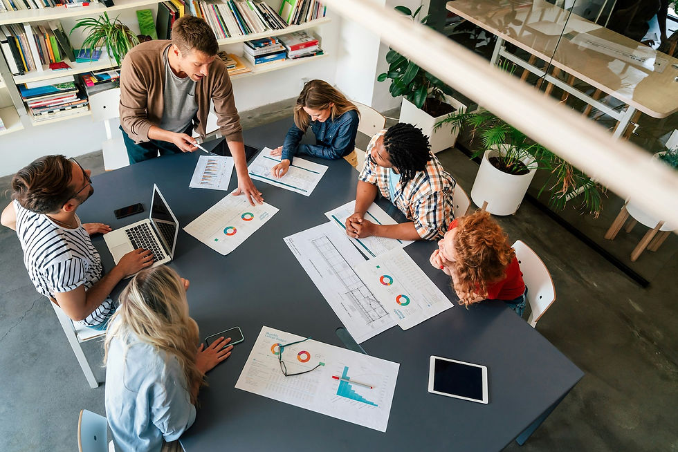 Six people in a meeting around a table with charts and laptops. Bright office, book-filled shelves, plants, collaborative atmosphere.