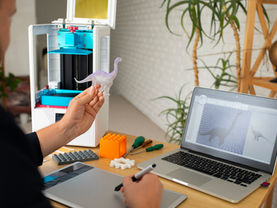 Ein BASE3D-Mitarbeiter arbeitet an einem industriellen 3D-Drucker, im Hintergrund ein gut organisierter Maschinenpark mit Filamentrollen, Monitoren und Prototypen.