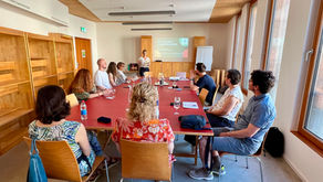A group of approximately ten people seated around a large red table in a bright, wood-panelled meeting room, attending a workshop on Nonviolent Communication. A facilitator stands at the front of the room next to a projected slide reading "Nonviolent Communication" and a flipchart.