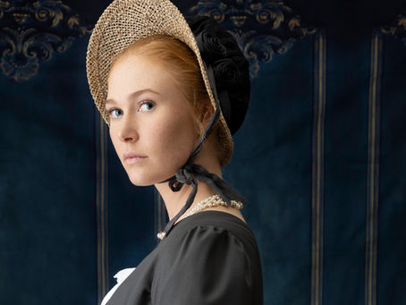 A woman dressed in Jane Austen–era Regency clothing, wearing a straw bonnet with black ribbons, a pearl necklace, and lace-trimmed gloves. She gazes sideways against an ornate dark blue Regency-style backdrop, evoking the atmosphere of early 19th-century England.