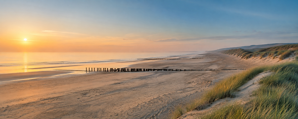 Wide landscape photo of a calm sandy beach at sunset with soft orange sky, gentle waves, and grassy dunes in the foreground