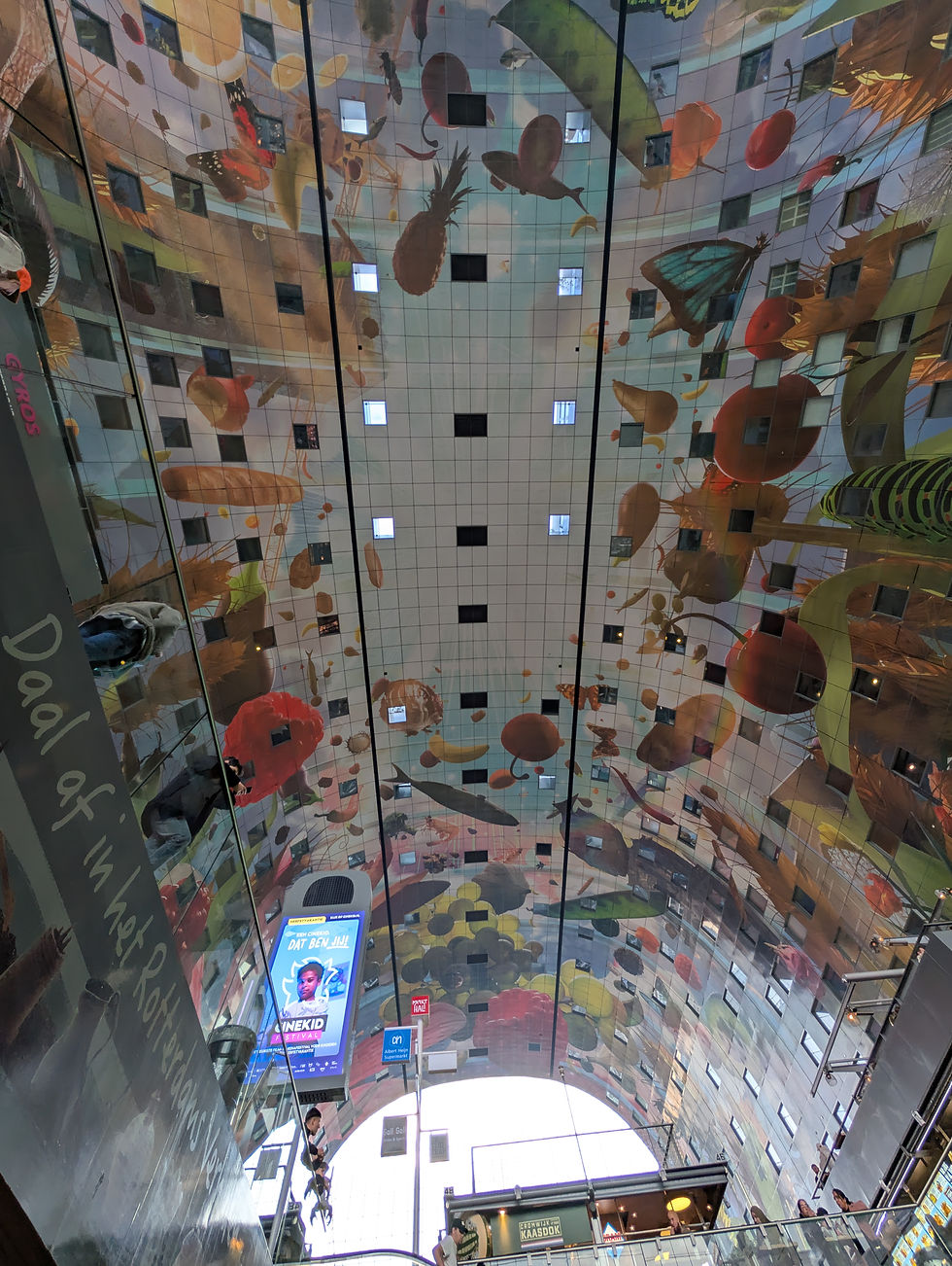 Colorful ceiling mural with fruits, vegetables, and butterflies at a market hall. People on escalators, digital signs visible. Text reads "Dat Ben Jij."