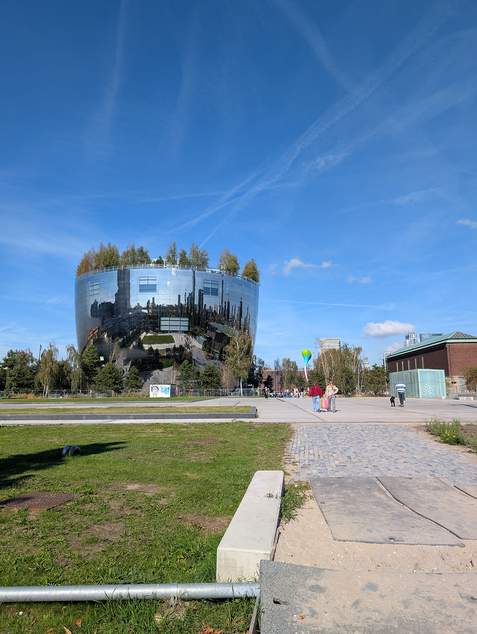 Reflective cylindrical building with rooftop trees under a clear blue sky. People walk on a path nearby. Vibrant and serene atmosphere.