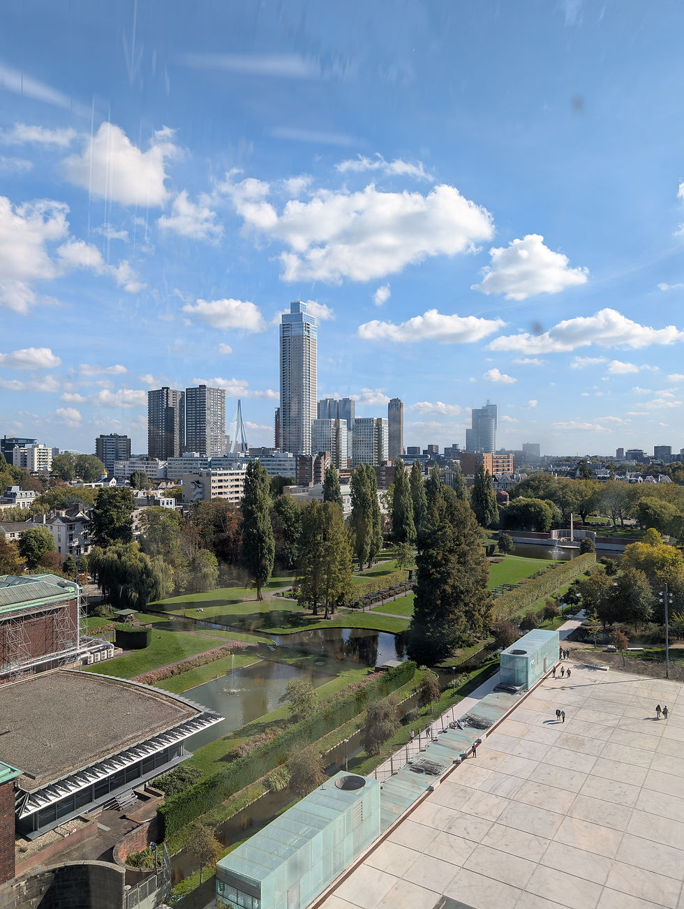 Cityscape with tall buildings under a blue sky with clouds. Park with trees and pathways below, people walking on a paved area.