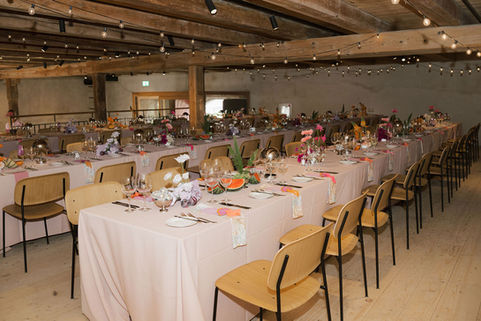Lange Hochzeitstafeln mit rosa Tischdecken und bunten Blumen unter Holzbalken