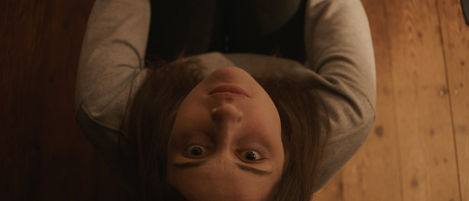 A woman looks up at the camera, from her position sat on the floor. She has brown hair and wears grey and black.