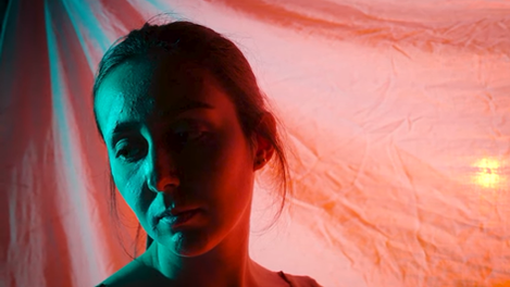 A woman with white paint streaked across her face looks sad, lit by blue light, with an orange background.
