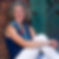 Photograph of Coach Jen Westra. Jen is sitting on the floor, leaning against a red brick wall. Jen is wearing a blue top with white pants. Jen is smiling and facing the camera.