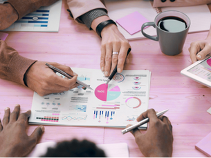 Overhead view of four people collaborating around a pink table, reviewing printed charts and graphs. Several hands hold pens and point to documents featuring pie charts, line graphs, and data visualisations.