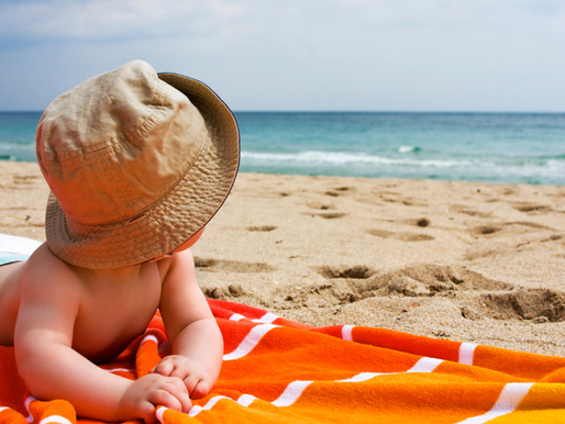 Baby in a nappy having tummy time on an orange and white stripe towel on the sand. Wearing brown hat looking out to sea.