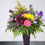 Thumbnail: A mix of pink, yellow and purple flowers and greenery in a tall dark purple vase on a white background.
