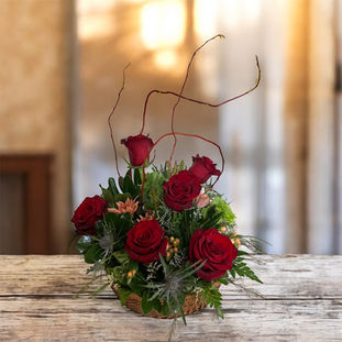 An arrangement of dark red roses, green and blue flowers in a basket with tall curly twigs.