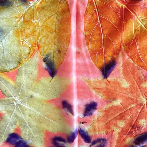 A small group gathered around a table at a craft workshop, carefully placing leaves and natural materials onto fabric to create ecoprint patterns, with tools and bundled textiles laid out nearby.