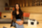 Simone, the CEO and founder of NIFT, is pictured sitting on a multi-tiered, light-colored wooden bench. She has long, dark brown hair and is wearing a black top, light blue jeans, and a brown and black patterned jacket. Her hands are clasped in her lap, and she is looking directly at the camera with a gentle smile. Behind her are several square pillows in shades of orange, brown, and blue, adding pops of color to the wooden backdrop. To her right, on the lower tier of the bench, sits a white shopping bag with black handles and the black word "NIFT" printed on the front. The setting appears to be a modern, possibly retail or event space, with vertical wooden slats visible in the background. The lighting is soft and warm.
