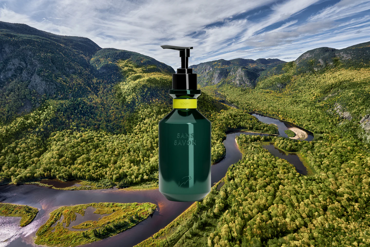 A dark green, cylindrical bottle of Sans Savon body wash with a black pump dispenser is digitally superimposed over a breathtaking aerial view of a lush, mountainous landscape with winding rivers. The bottle features a vibrant yellow-green band around its neck with the words "SANS SAVON" vertically printed on its front. The background showcases a vast expanse of green forests covering rolling hills and mountains, with several dark rivers snaking through the valleys. The sky above is partly cloudy with patches of blue. The juxtaposition of the product with this pristine natural environment likely aims to emphasize the brand's connection to nature or its pure ingredients.