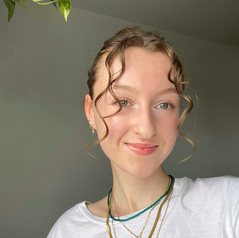 A softly lit selfie of Ella, a nail influencer, shows her smiling gently at the camera. Her light brown, wavy hair is styled with loose curls around her face. She has fair skin and light eyes. She is wearing a white t-shirt with a red graphic and several layered necklaces in gold and green beads. A green and white variegated plant is visible in the upper left corner. The background is a plain, light grey wall.