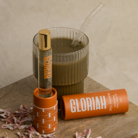 A glass perfume bottle with a gold cap is positioned in an orange cylindrical holder with a repeating geometric pattern. Next to it is a matching orange tube labeled "Gloriah Hey, Sunshine Perfuming Pulse Point Oil". A glass with a green-brown drink and a clear straw is visible in the background. Scattered pink and white petals are around the base of the perfume holder. The items are placed on a light-colored wooden surface with a visible grain.