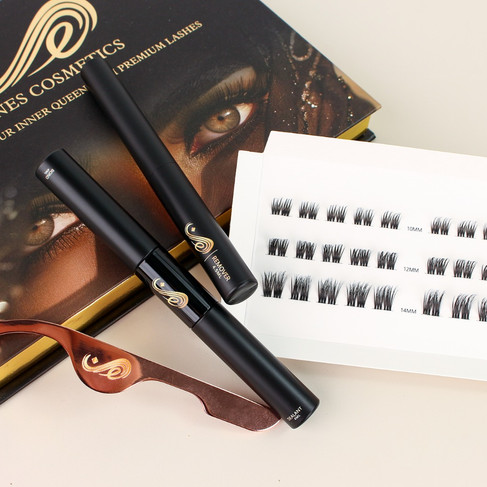 An assortment of Inês Cosmetics lash application products is arranged on a light grey surface. The main product box, black with gold accents and an image of a woman's eye with dramatic lashes, is partially visible in the background. In the foreground, there are several strips of individual false lash clusters adhered to a white card, with varying lengths indicated by printed numbers (10mm, 12mm, 13mm, 14mm). Below the lashes, a brown lash applicator tool with a distinctive gold "I" logo cutout is positioned. Two sleek black tubes, likely containing lash adhesive and sealant, also feature the gold "I" logo and the brand name. The overall presentation is stylish and highlights the tools and lashes needed for a complete application.
