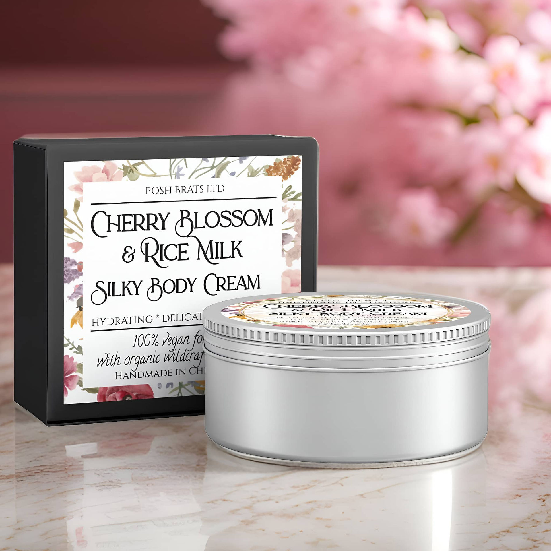 A silver tin of "Posh Brats Cherry Blossom & Rice Milk Silky Body Cream" sits on a light-colored marble surface in front of its black and floral-decorated box. The tin has a screw-top lid with a label matching the box. The background is softly blurred, showing pink cherry blossom branches. The scene is well-lit, highlighting the product and its packaging.