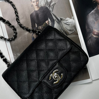 A black Chanel Mini Square bag is elegantly placed on top of an open fashion magazine. The bag is made of textured black leather with the iconic diamond quilting pattern. It features the silver Chanel double "C" logo on the front flap and a silver chain strap interwoven with black leather. The magazine pages underneath showcase high-fashion photography, with models wearing sophisticated attire. The background is a light grey surface. The lighting is soft and diffused, highlighting the texture of the bag and the glossy pages of the magazine.