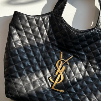 A close-up shot showcases a black Saint Laurent Icare Quilted Top Handle Tote bag resting on a light-colored surface, likely concrete or stone, with shadows cast across it. The tote is made of quilted black leather in a diamond pattern and features the iconic gold-toned YSL logo prominently displayed in the center. The bag has two top handles and a spacious, open-top design. The texture of the quilted leather and the shiny gold logo are clearly visible, emphasizing the luxury and design of the handbag.