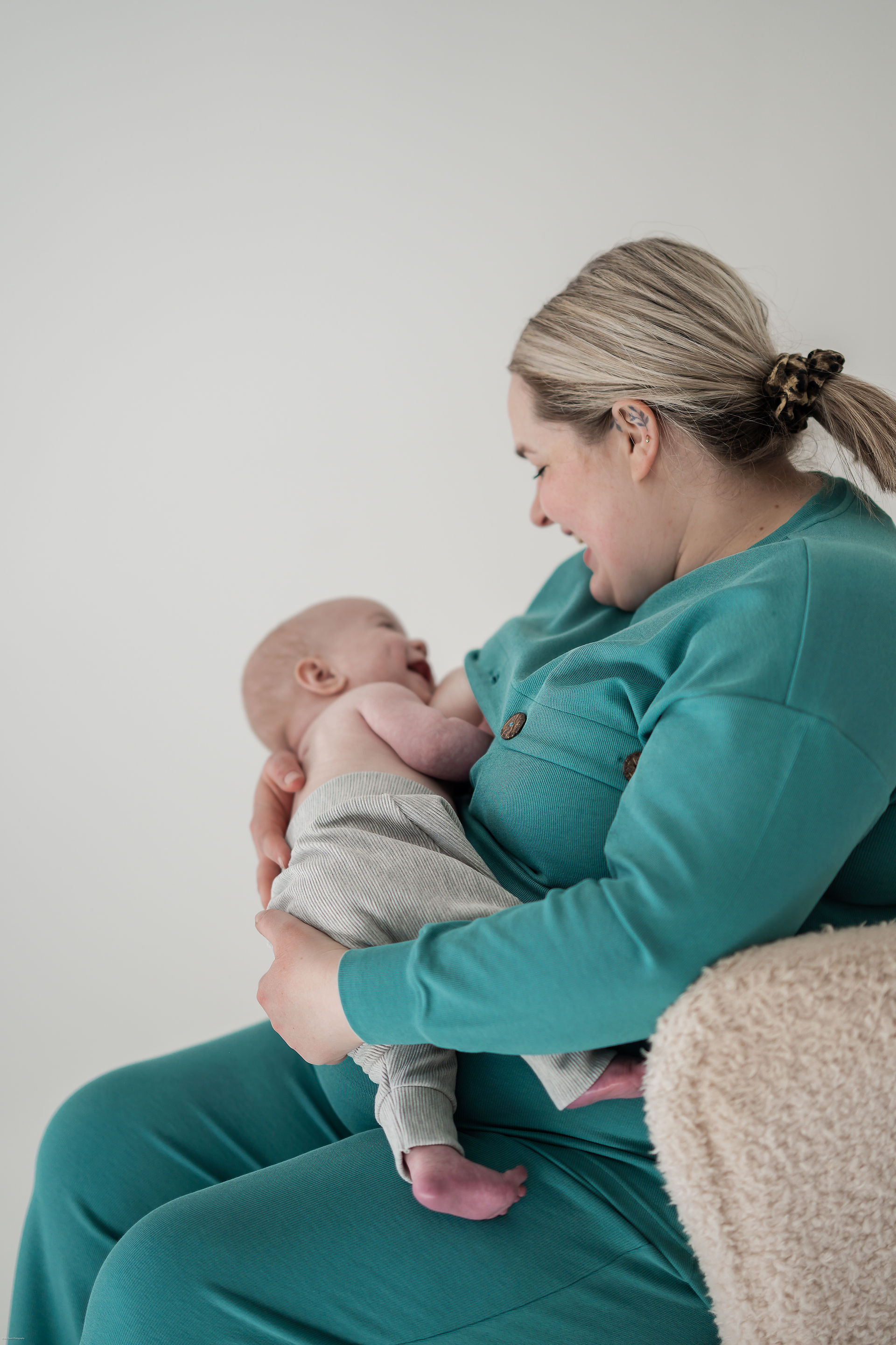 A woman with blonde hair pulled back in a ponytail secured with a brown leopard-print scrunchie is cradling a baby in her arms. The woman is wearing a teal, long-sleeved top with buttons on the shoulder and matching teal trousers. She is looking down at the baby with a loving smile. The baby, who is mostly bald, is wearing light grey ribbed leggings and is looking up at the woman with a happy expression, reaching out with its hands. They are seated on a beige, textured surface, possibly a chair or cushion, against a plain white background. The lighting is soft and even, emphasizing the tender interaction between the mother and child.