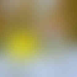 A yellow, disc-shaped "Chamomile Aromatherapy Shower & Bath Steamer" sits next to its gold foil wrapper on a textured, light-colored surface. The wrapper has a colorful label featuring a sunflower, the word "Chamomile," and text indicating it contains pure chamomile essential oil and is an aromatherapy steamer. The background is blurred, showing out-of-focus yellow and green foliage.