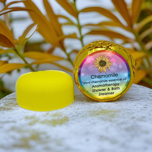 A yellow, disc-shaped "Chamomile Aromatherapy Shower & Bath Steamer" sits next to its gold foil wrapper on a textured, light-colored surface. The wrapper has a colorful label featuring a sunflower, the word "Chamomile," and text indicating it contains pure chamomile essential oil and is an aromatherapy steamer. The background is blurred, showing out-of-focus yellow and green foliage.