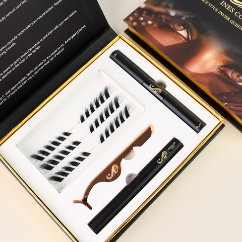 An open Inês Cosmetics lash application kit is presented from an overhead perspective. The kit is contained within a gold-trimmed black box, the lid of which is partially visible at the top right, featuring an image of a woman's eye with prominent lashes and the "Inês Cosmetics" logo. Inside the open portion of the box, various lash application tools and lashes are neatly arranged in a white insert. On the left side, illustrated instructions for applying the lashes are printed in black text. The kit includes several strips of individual false lash clusters, a brown lash applicator tool, and two black tubes, likely containing lash adhesive or sealant. The overall presentation is clean and organized, emphasizing the products and the step-by-step guide for use.