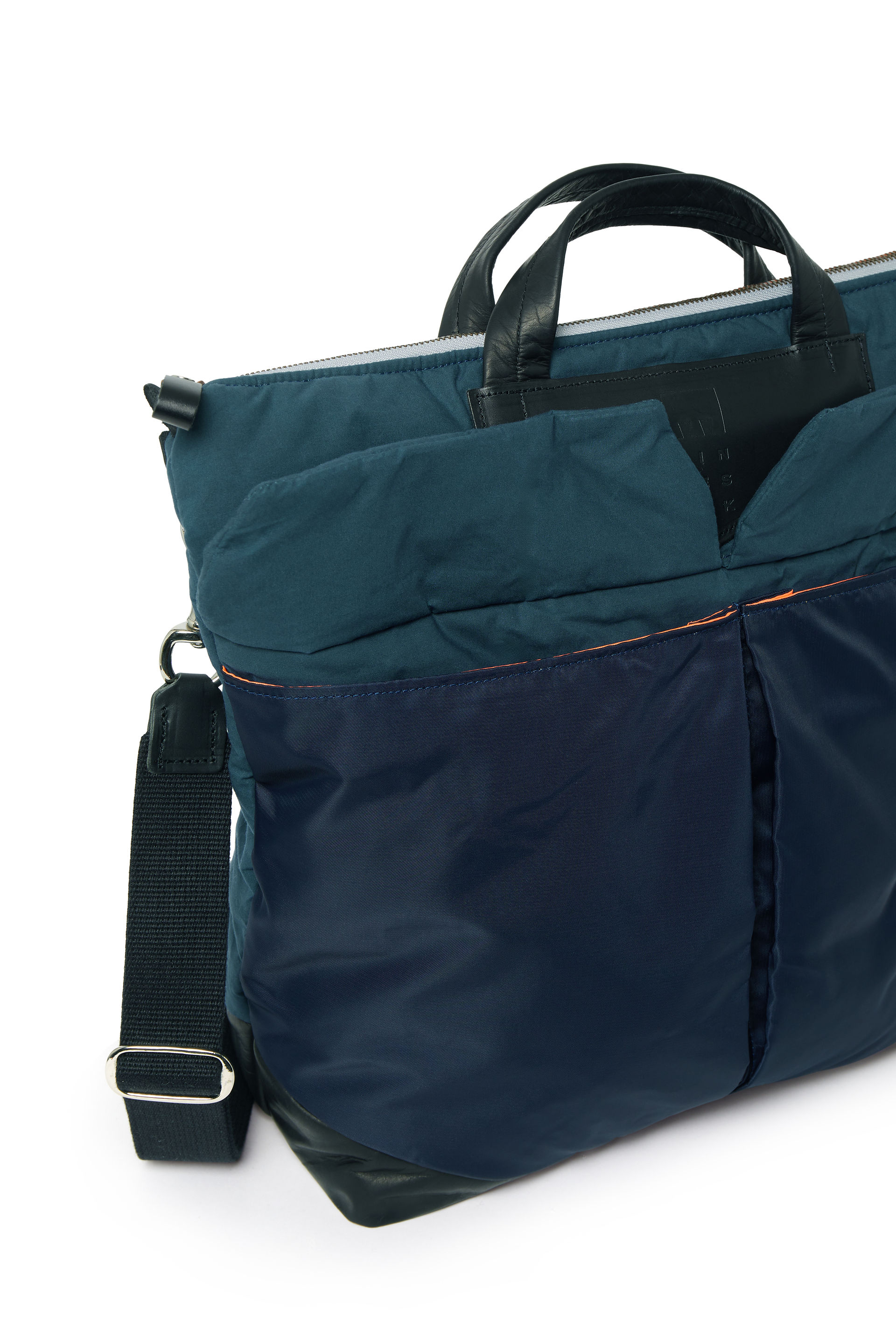 A close-up shot focuses on a stylish, multi-toned blue and black tote bag against a bright white background. The bag features a combination of shiny and matte nylon fabrics in shades of deep teal and navy blue. It has black leather top handles and a detachable black nylon shoulder strap with silver hardware. An orange accent line is visible along the edge of one of the front pockets. The bag appears to be well-constructed with multiple compartments and a sleek, modern design. The lighting is even, highlighting the different textures and colors of the bag.