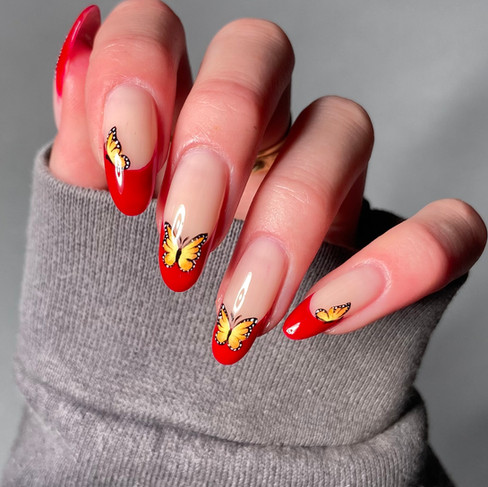 A close-up of a hand displays long, oval-shaped nails with a vibrant red French tip. Each red tip is decorated with a detailed yellow and black butterfly decal. The hand rests on a grey sweater sleeve. The lighting is bright, showcasing the intricate nail art.